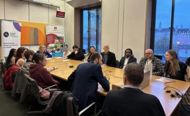 A group of people are seated around a large wooden conference table in a formal meeting room with tall windows. There are banners in the background. There is a panel of people including members of parliament engaged in conversation, with some taking notes or listening attentively.
